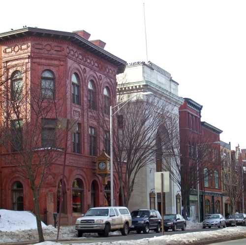 Historic buildings line a snowy street, showcasing red brick architecture and a prominent archway in the background.