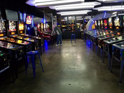 A dimly lit arcade with rows of pinball machines and a person playing at one of the machines.