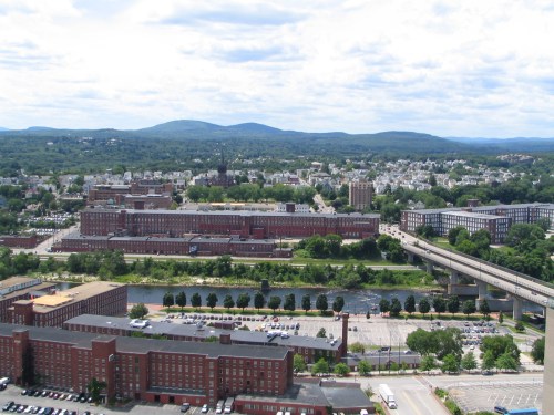 Aerial view of a city with brick buildings, a river, and green hills in the background under a partly cloudy sky.
