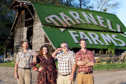 Four people stand in front of a rustic barn with "Darnell Farms" painted on the roof, smiling and posing together.