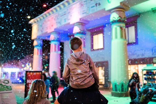 A child on an adult's shoulders watches colorful lights and falling snow at a festive outdoor event.