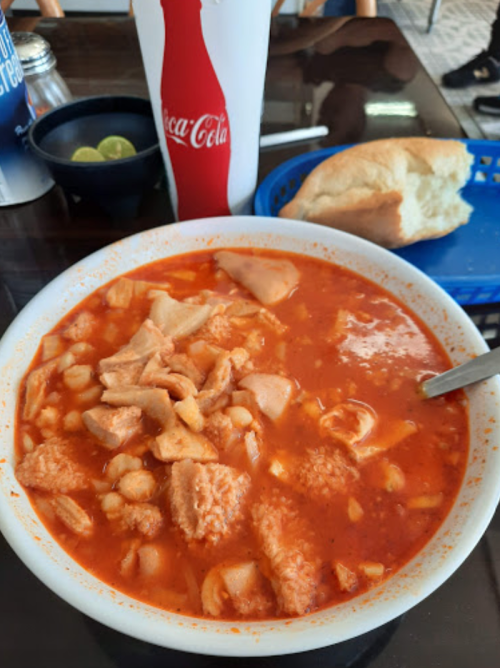 A bowl of red soup with chicken, hominy, and crispy toppings, served with a bread roll and a soda.
