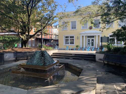 A sunny park scene featuring a fountain, benches, and a yellow building in the background surrounded by trees.