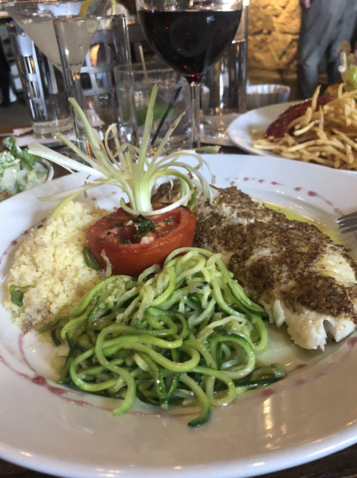A plate featuring seasoned fish, zucchini noodles, couscous, a baked tomato, and a glass of red wine in the background.