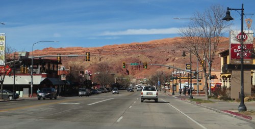A street view with shops and traffic lights, framed by red rock formations under a blue sky.