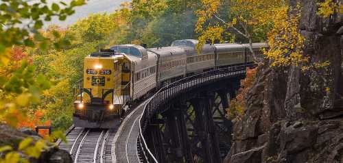 A yellow train crosses a bridge surrounded by vibrant autumn foliage and rocky terrain.