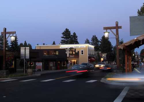 A quiet street scene at dusk, featuring shops, street lamps, and moving cars in a small town.