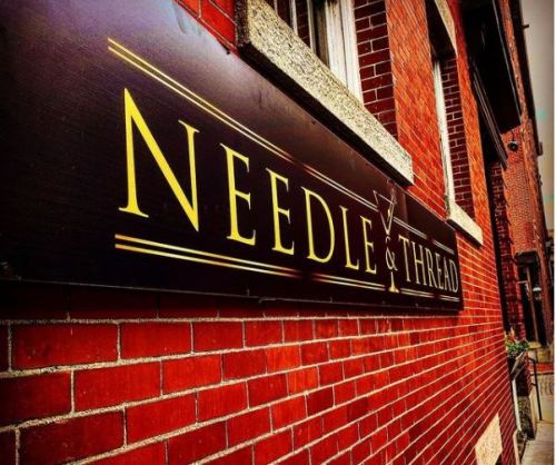 Sign for "Needle & Thread" on a brick wall, featuring elegant lettering in gold against a dark background.