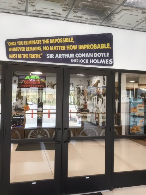 Sign above glass doors featuring a quote from Sherlock Holmes by Sir Arthur Conan Doyle. Interior displays visible.