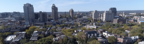 Aerial view of a city skyline with tall buildings and green trees, under a clear blue sky.