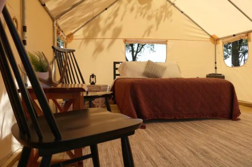 Cozy glamping tent interior with a bed, chairs, and warm lighting, surrounded by nature.