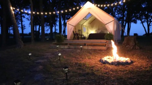 A cozy tent at night, illuminated by string lights, with a campfire and lanterns in a wooded area.