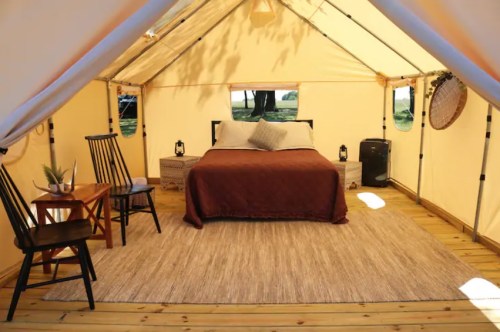 Cozy glamping tent interior with a bed, chairs, table, and decorative elements on a wooden floor.