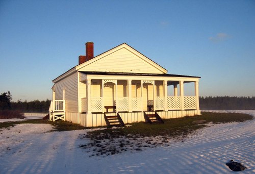 A white house with a porch sits on snowy ground under a clear blue sky, surrounded by trees in the distance.