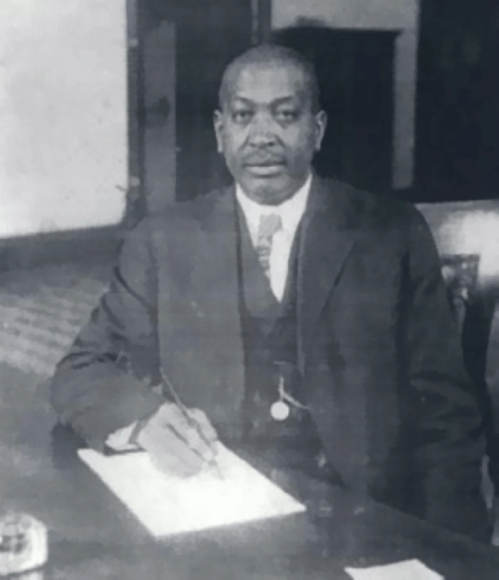 A historical black-and-white photo of a man in a suit, seated at a table, writing on a piece of paper.