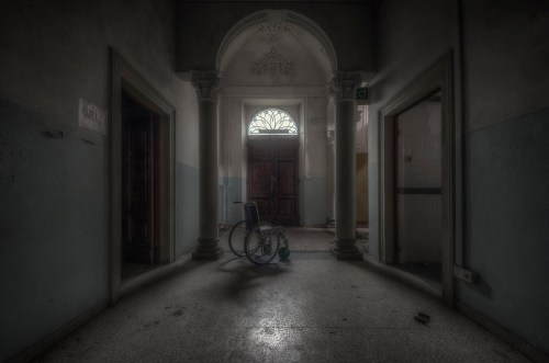 An empty, dimly lit hallway with a lone wheelchair in the center, framed by ornate architecture and closed doors.