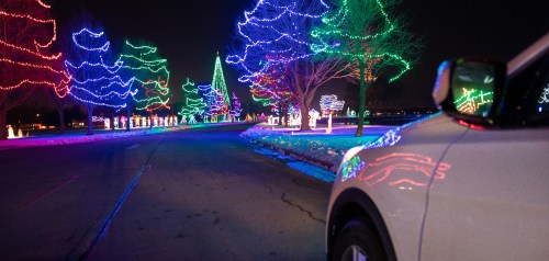 A car parked on a road lined with colorful holiday lights and illuminated trees at night.