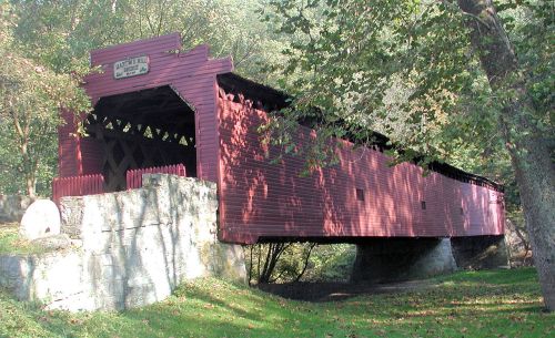 A red covered bridge spans a small creek, surrounded by trees and greenery.