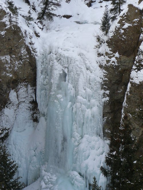 A frozen waterfall surrounded by snow-covered rocks and evergreen trees in a winter landscape.