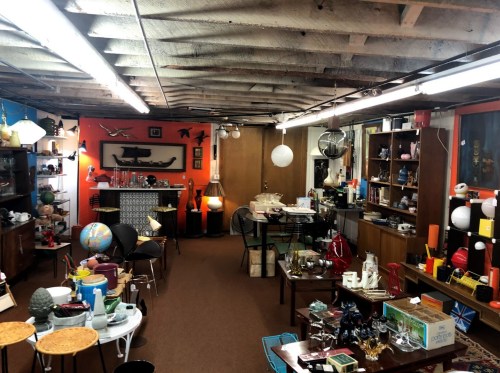 A vintage shop filled with various antiques, furniture, and decor under a rustic ceiling with bright overhead lights.