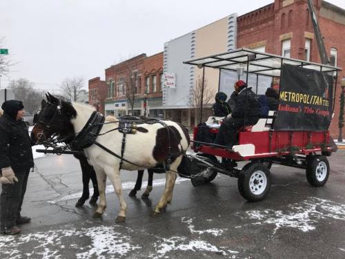 A horse-drawn carriage waits on a snowy street, with two passengers and a driver in warm clothing. Historic buildings in the background.