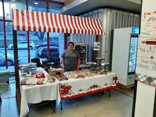 A vendor stands behind a table filled with various baked goods under a striped canopy in a market setting.