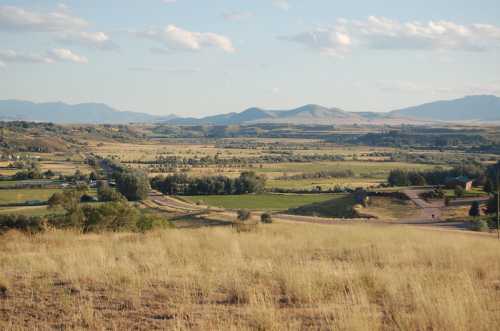 A scenic view of rolling hills and valleys under a blue sky, with fields and a winding road in the foreground.