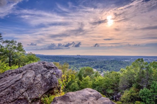 A scenic view from a rocky overlook, featuring lush green trees and a cloudy sky with the sun setting in the background.