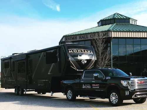 A black RV labeled "Montana" hitched to a GMC truck, parked in front of a building with a green roof.