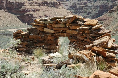 Stone archway made of stacked rocks, set against a backdrop of rugged canyon terrain and sparse vegetation.