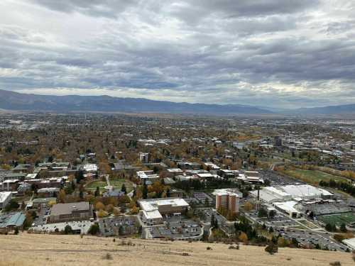 Aerial view of a city surrounded by mountains, featuring a mix of buildings and autumn-colored trees under a cloudy sky.