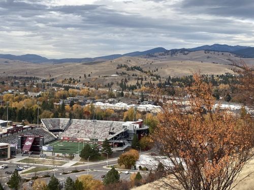 Aerial view of a stadium surrounded by autumn trees and rolling hills under a cloudy sky.