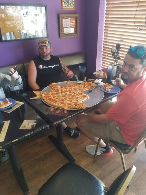Two men sit at a table in a restaurant, enjoying a large pizza and side dishes. The walls are decorated with framed photos.