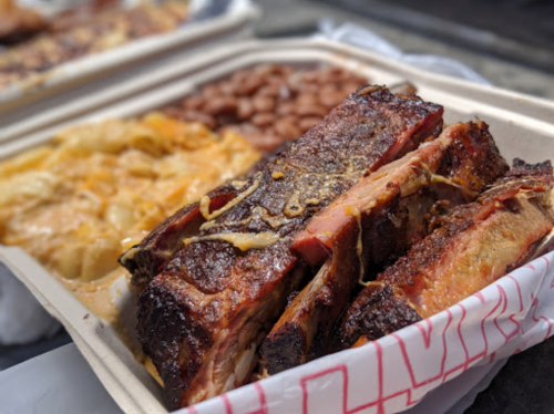 A close-up of smoked ribs with a side of baked beans and cheesy potatoes in takeout containers.