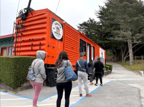 A group of people stands in line outside an orange food truck surrounded by greenery.
