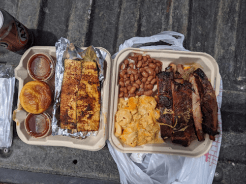 Two takeout containers filled with ribs, macaroni and cheese, baked beans, cornbread, and sauces on a textured surface.