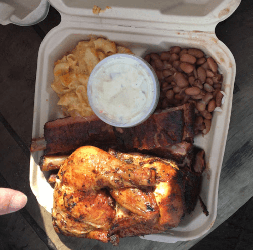 A takeout container with grilled chicken, ribs, macaroni and cheese, baked beans, and a side of coleslaw.