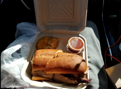 A takeout container with a sandwich, a side of sauce, and a small portion of food, resting on a person's lap.
