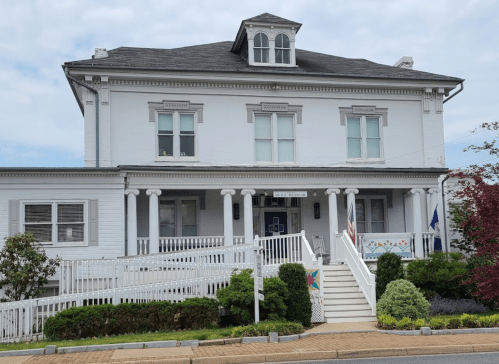 A large white house with a porch, surrounded by greenery and a walkway, under a cloudy sky.