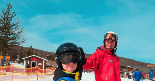 Two skiers on a snowy slope, one wearing a blue jacket and helmet, the other in a red jacket, smiling at the camera.