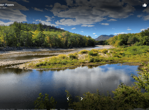 A serene river landscape with lush green trees and mountains under a blue sky with fluffy clouds.