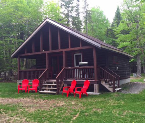 A cozy wooden cabin surrounded by trees, featuring red chairs on the lawn and a welcoming porch.