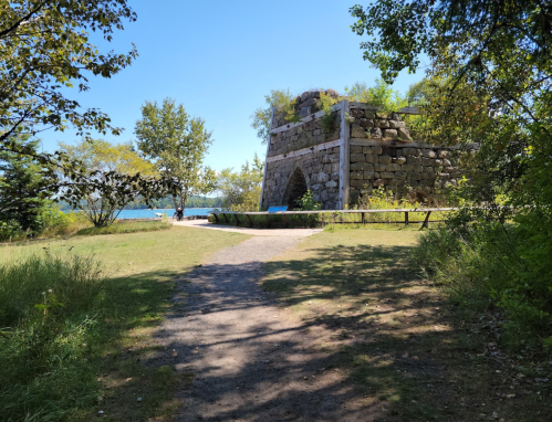 A stone structure surrounded by trees, with a path leading to a waterfront under a clear blue sky.