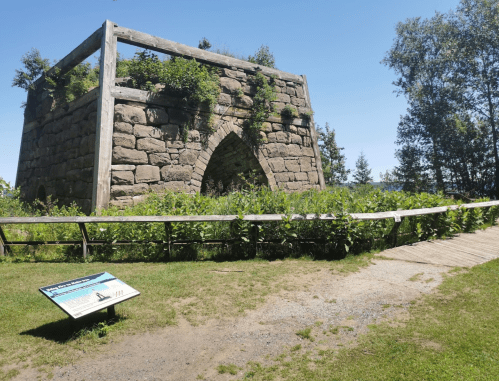 Ruins of a stone structure covered in greenery, with a wooden walkway and informational sign in the foreground.