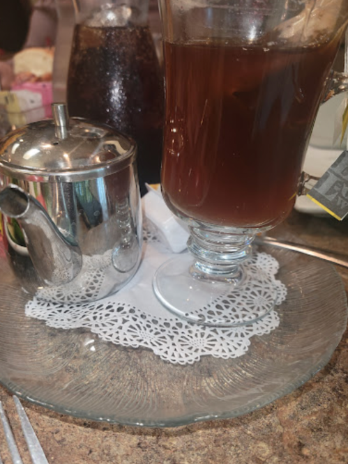 A glass of dark tea on a lace doily, next to a silver teapot and a bottle of soda in the background.