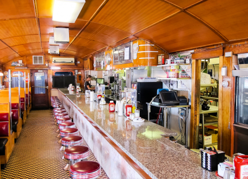 A vintage diner interior featuring a long counter with stools, checkered floor, and a kitchen area in the background.