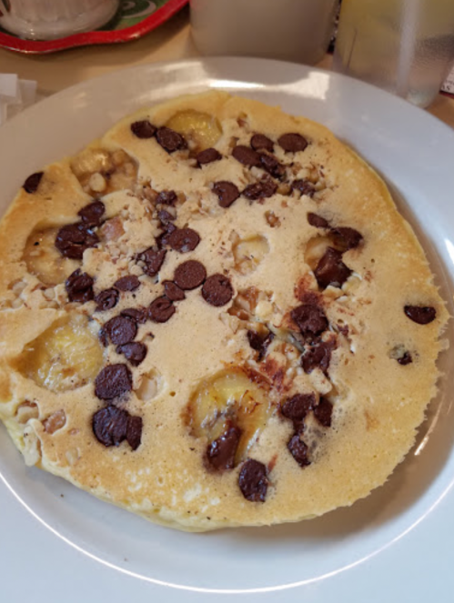 A fluffy pancake topped with banana slices, chocolate chips, and chopped nuts, served on a white plate.