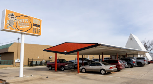 Exterior of Patty Wagon restaurant with a sign, cars parked under a canopy, and a triangular building in the background.
