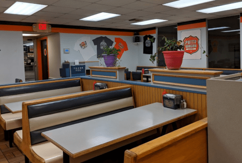 Interior of a casual dining restaurant with booths, tables, and decorative plants. Walls feature t-shirts and signage.