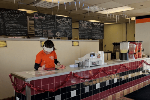 A staff member in an orange shirt takes orders at a counter decorated with festive garlands and menu boards.
